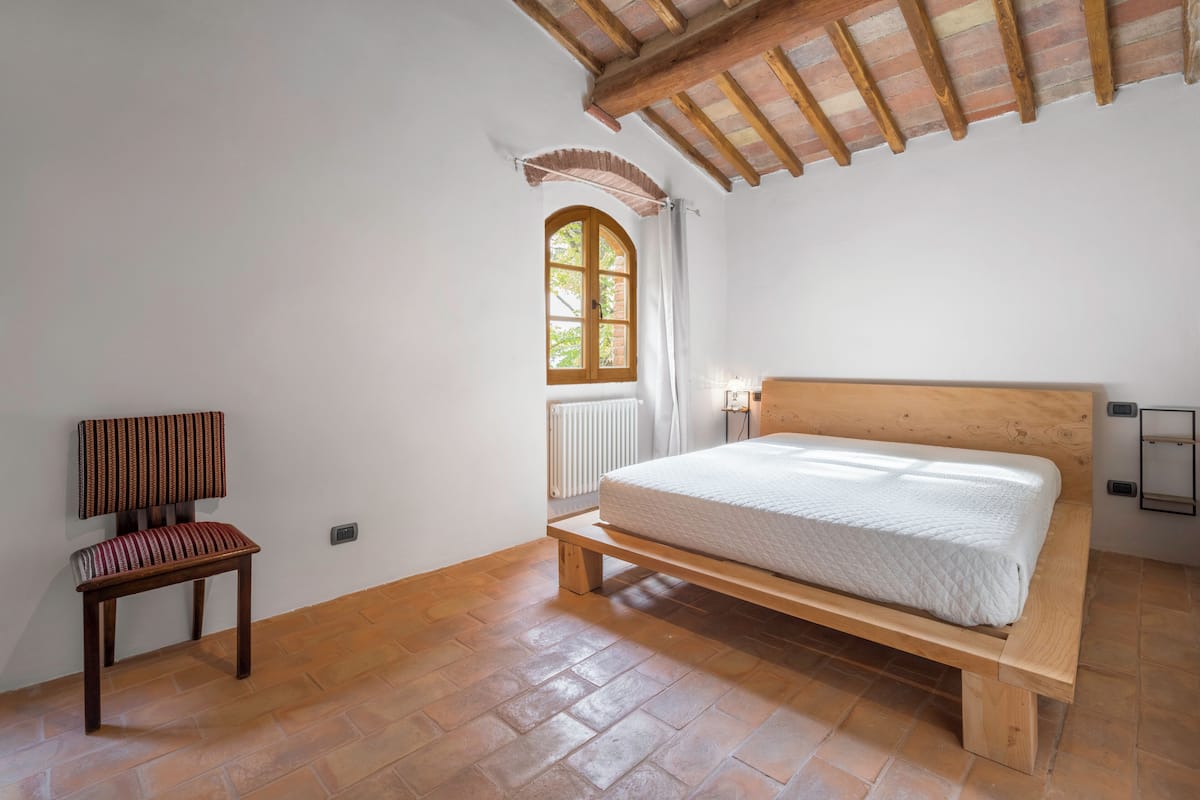 Ein Schlafzimmer mit einem hölzernen Bett, einem Sessel, einem Fenster mit Vorhängen und einer Heizung.