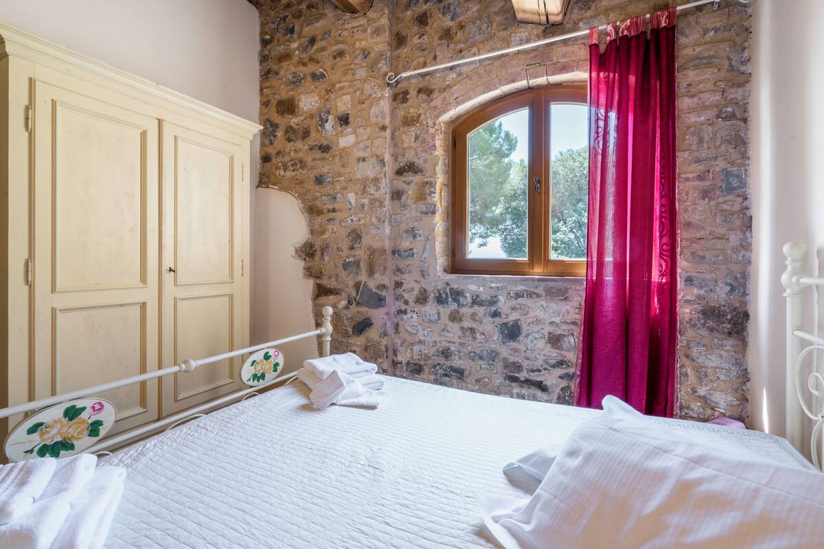 Ein Schlafzimmer mit Steinwand, einem großen Fenster mit roten Vorhängen, einem Bett mit weißen Leinen und einem Holzschrank.