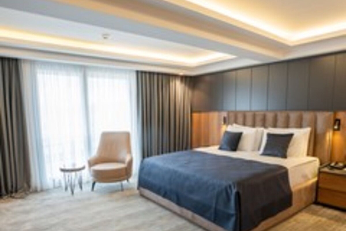 A modern hotel room with a large bed, a chair, a nightstand, and a view of the outdoors through the window.