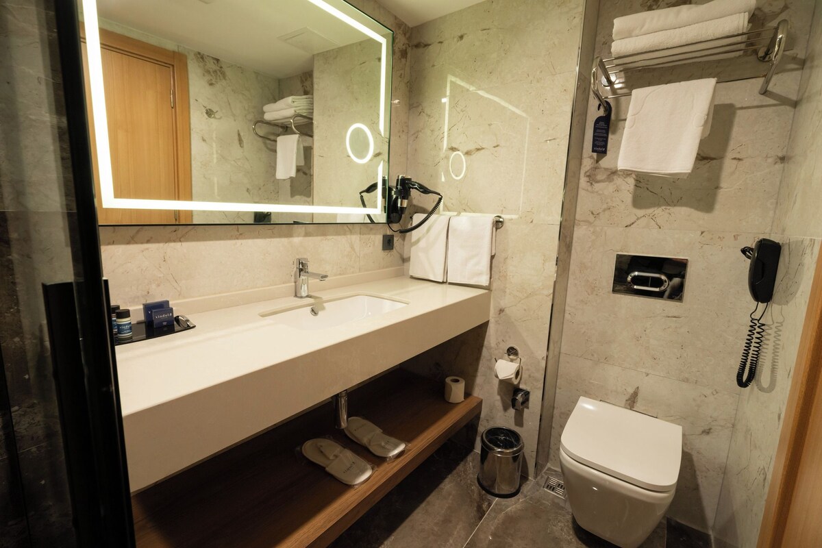 A modern bathroom with a marble sink, a large mirror, a hairdryer, a toilet, and a phone.