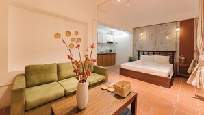 A modern hotel room with a green sofa, a wooden coffee table, a vase with flowers, a bed with a wooden headboard, and a kitchen area in the background. - Ha Noi Memory Apartel (Hanoi)