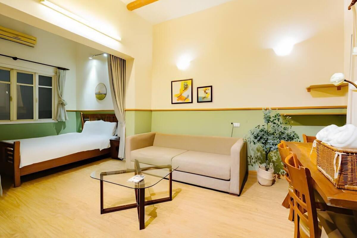 A hotel room with a bed, a sofa, a glass coffee table, a dining table with chairs, a potted plant, and two framed pictures on the wall.
