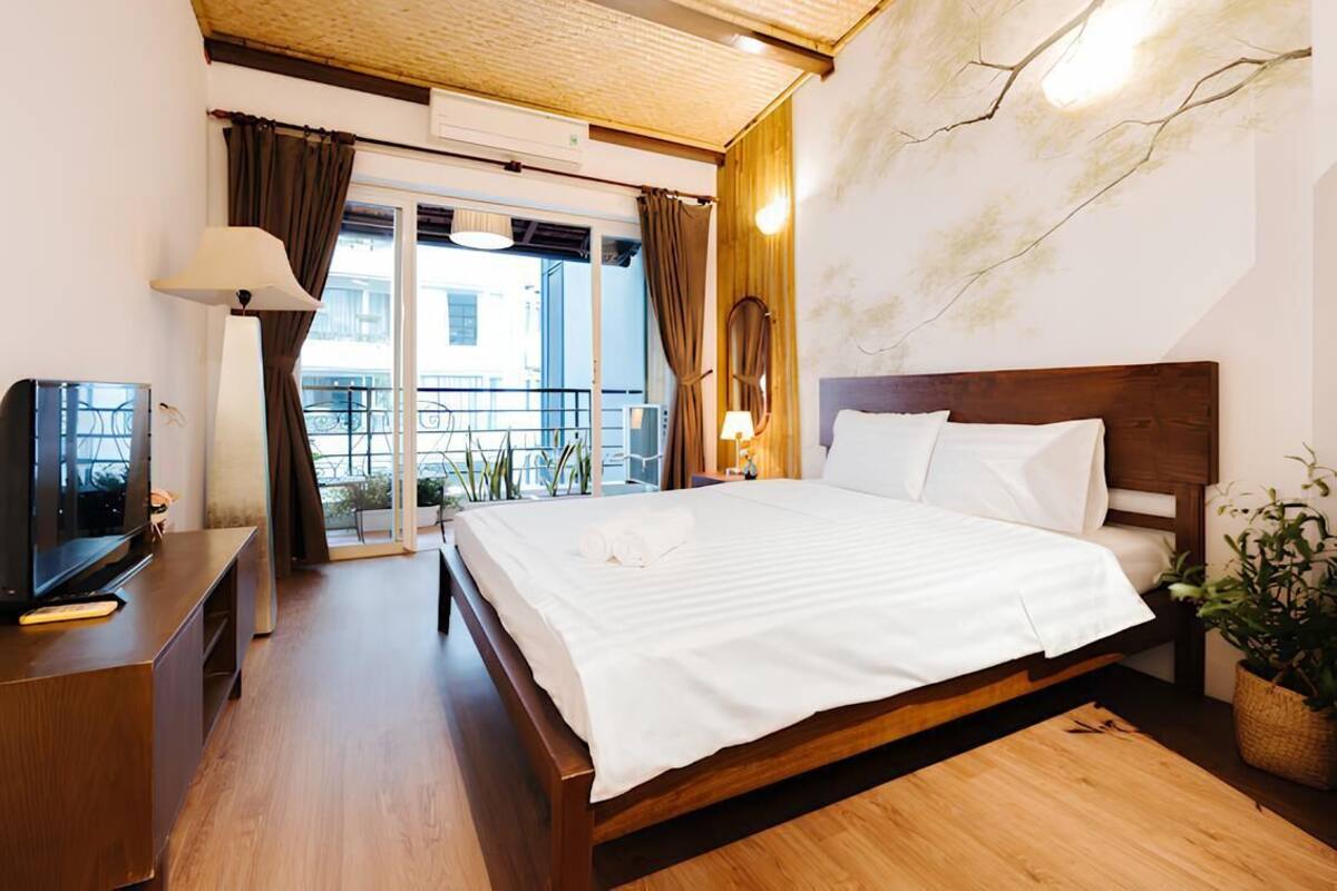 A hotel room with a wooden bed, a TV on a wooden cabinet, a balcony with curtains, and a view of buildings outside.