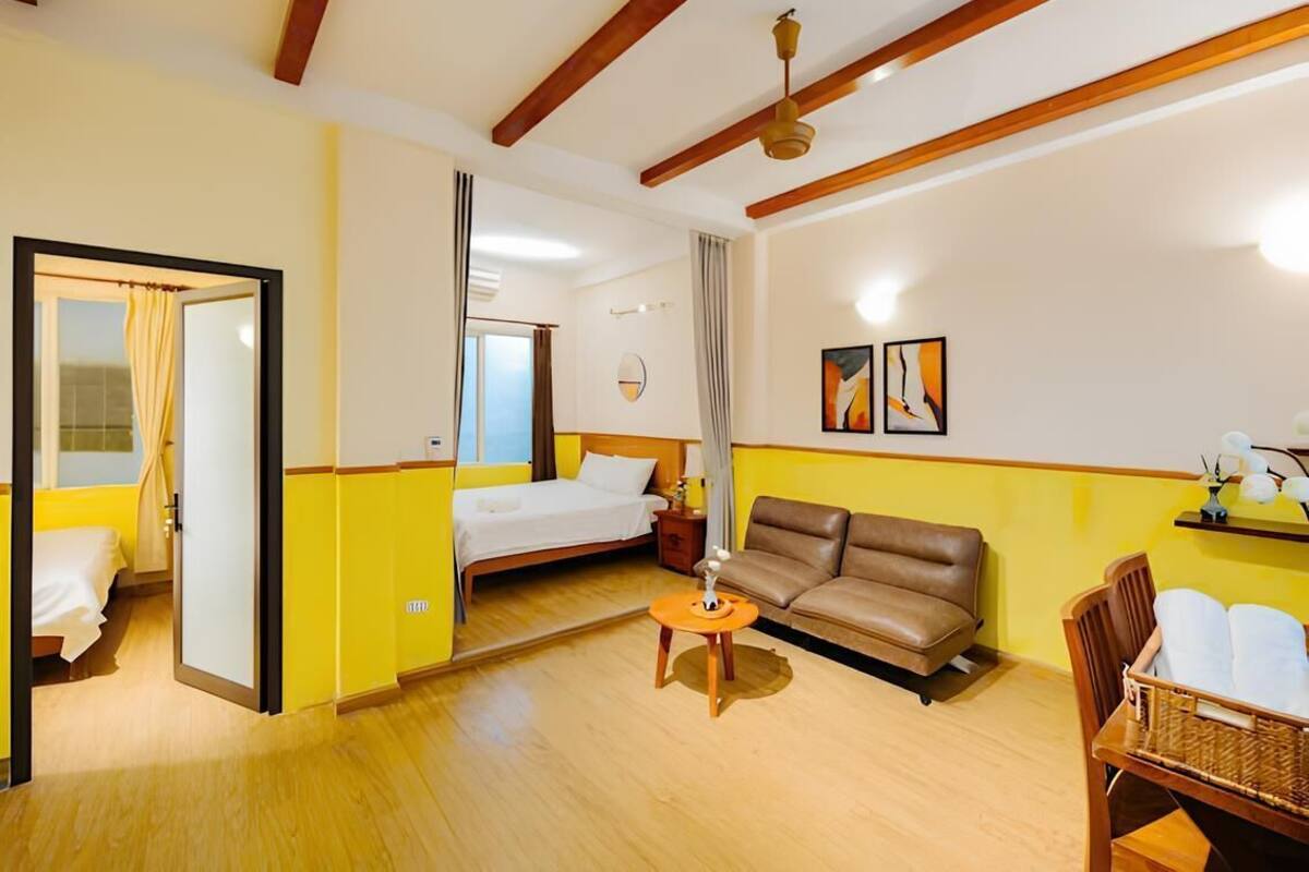 A hotel room with a bed, a sofa, a small table, and a chair. There is a window with curtains, a ceiling fan, and a wall-mounted shelf with items.