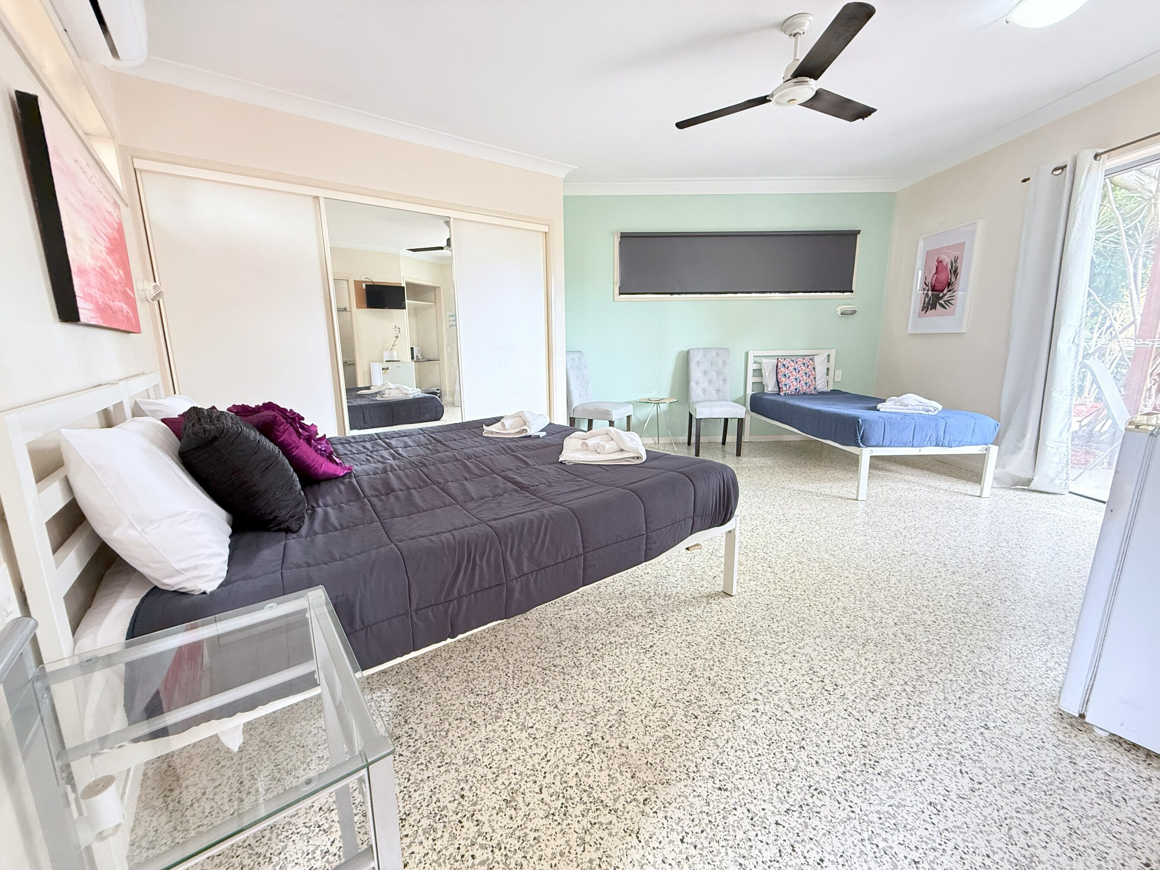 A bedroom with a bed, a chair, a small table, a ceiling fan, and a large window.