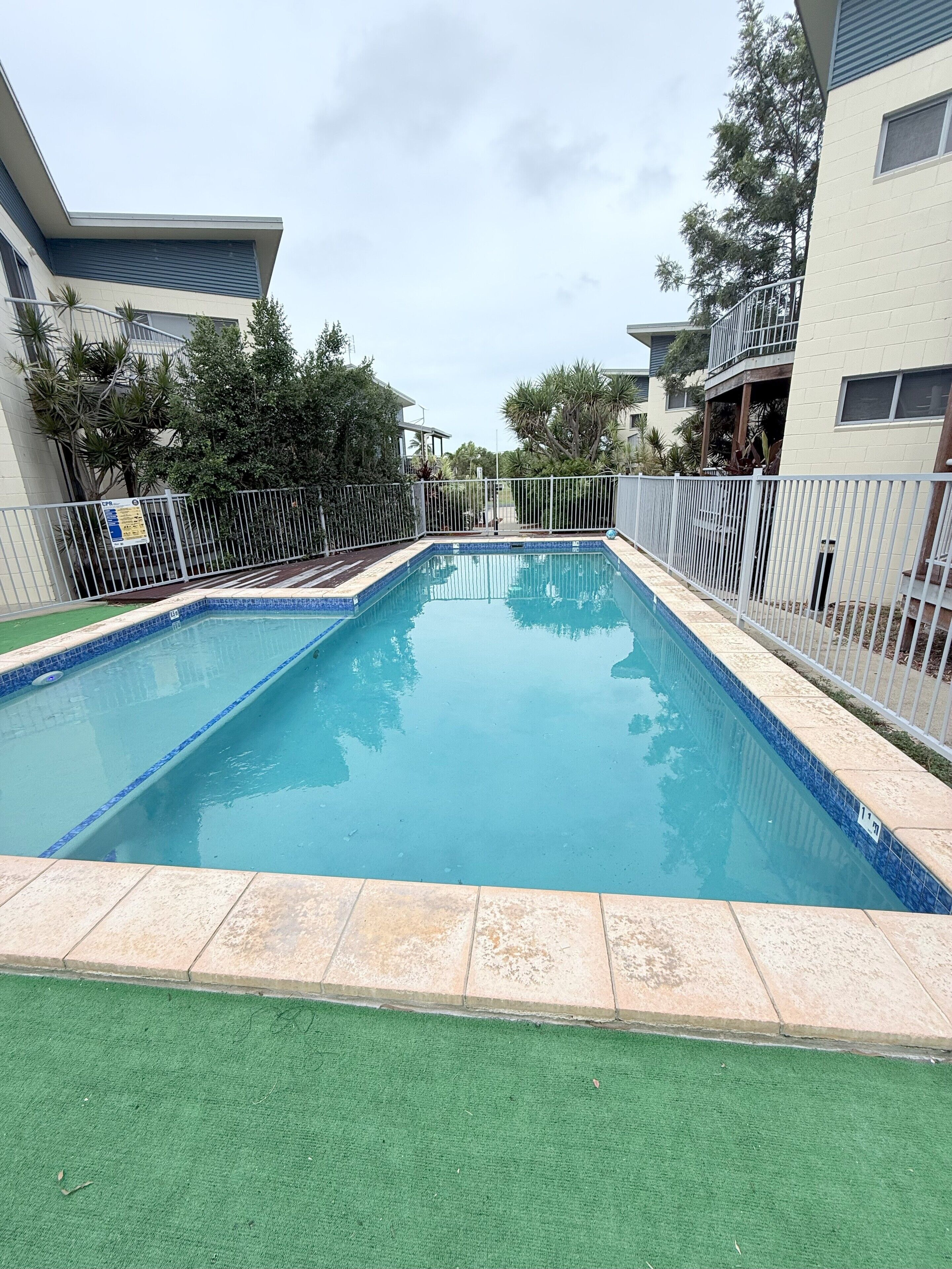 A rectangular swimming pool with a tiled edge, surrounded by a white fence and a green artificial turf area in the foreground.
