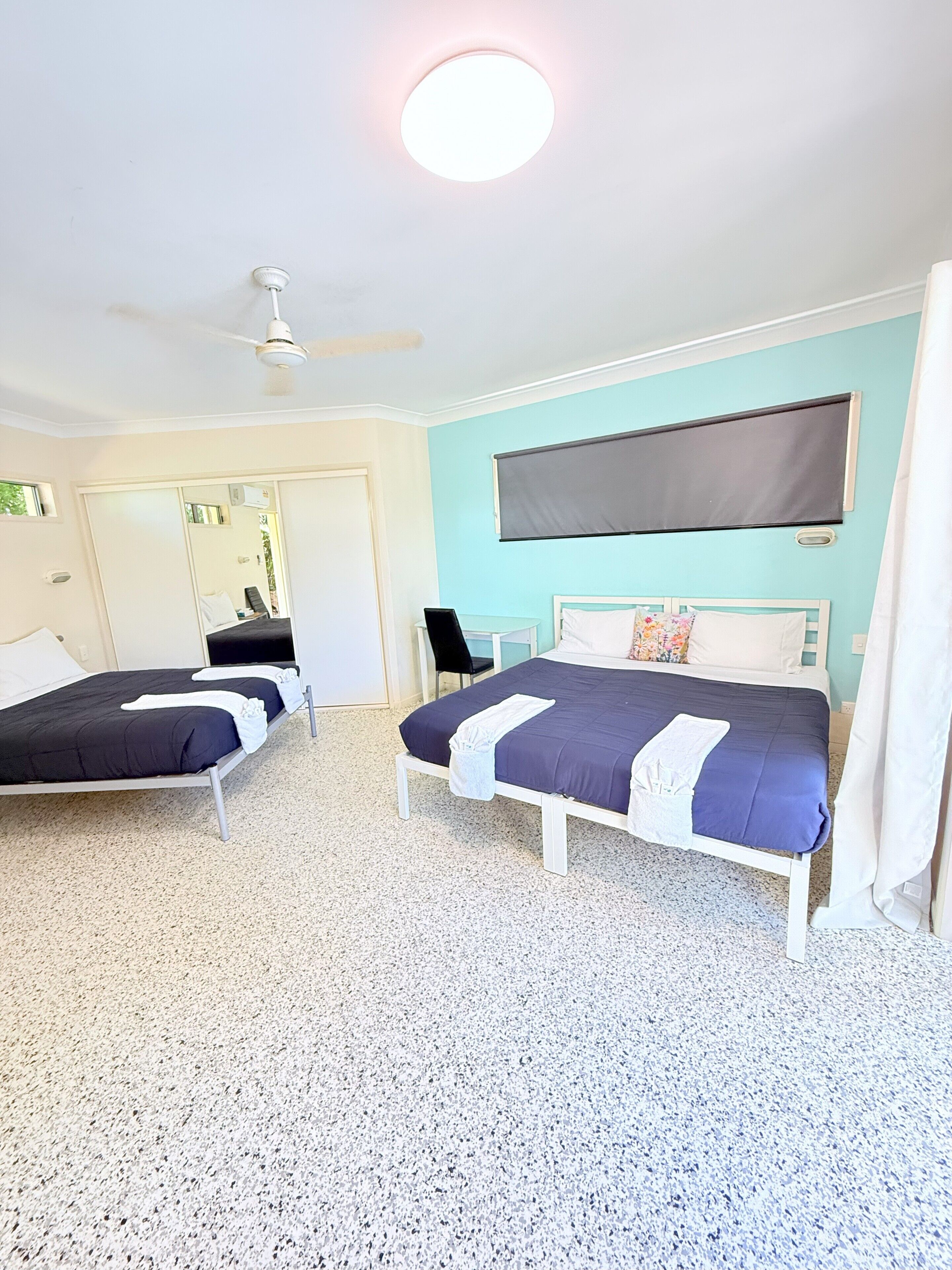 A hotel room with two beds, a ceiling fan, a large window with a blind, and a small desk with a chair.