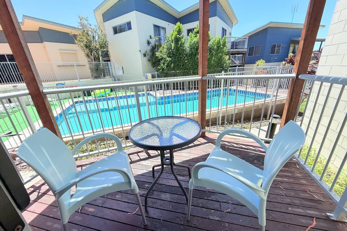 Eine Holzterrasse mit einem runden Tisch und zwei weißen Stühlen mit Blick auf einen Swimmingpool und Wohnhäuser.