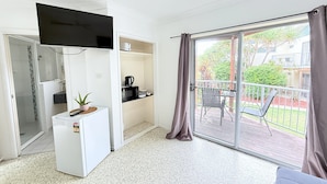 A compact living space with a kitchenette, a flat-screen TV, a small refrigerator, a dining area with a table and chairs, and a sliding glass door leading to an outdoor patio with a view of greenery and a building. - Emu Park Beach Resort (Emu Park)