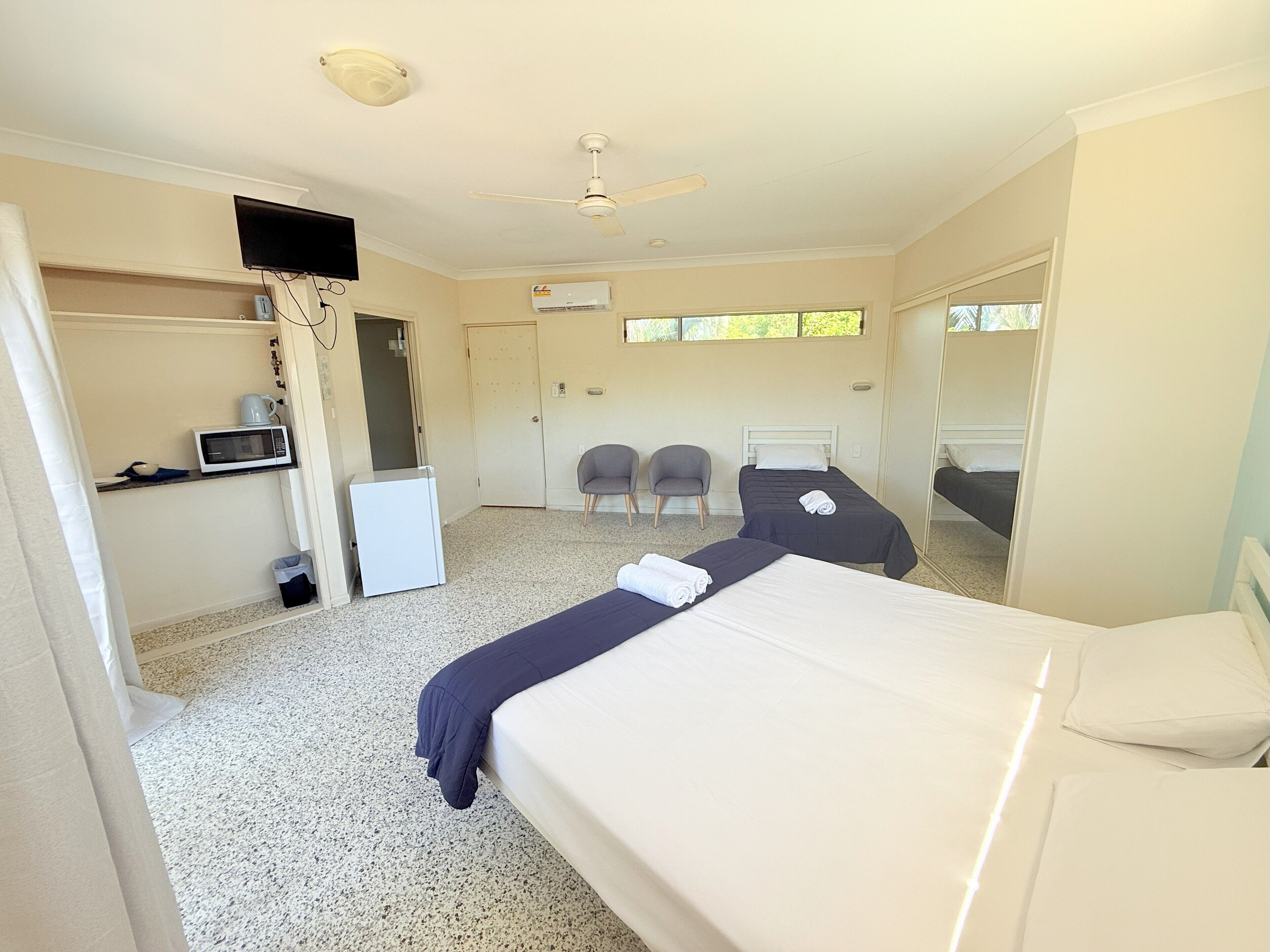 A hotel room with a bed, a desk with a computer, a microwave, a refrigerator, two chairs, and a ceiling fan.