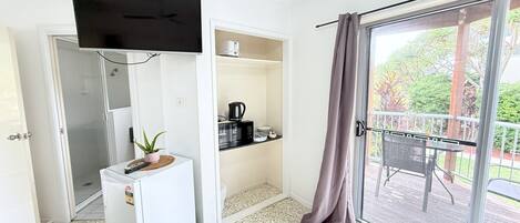 A compact living space with a kitchenette, a small fridge, a TV mounted on the wall, and a sliding glass door leading to an outdoor area with a table and chairs.