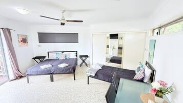 Une chambre avec deux lits, un ventilateur de plafond, une télévision, une petite table avec une plante et une fenêtre avec des rideaux.