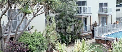 A modern building with multiple balconies, a swimming pool, and a variety of tropical plants.