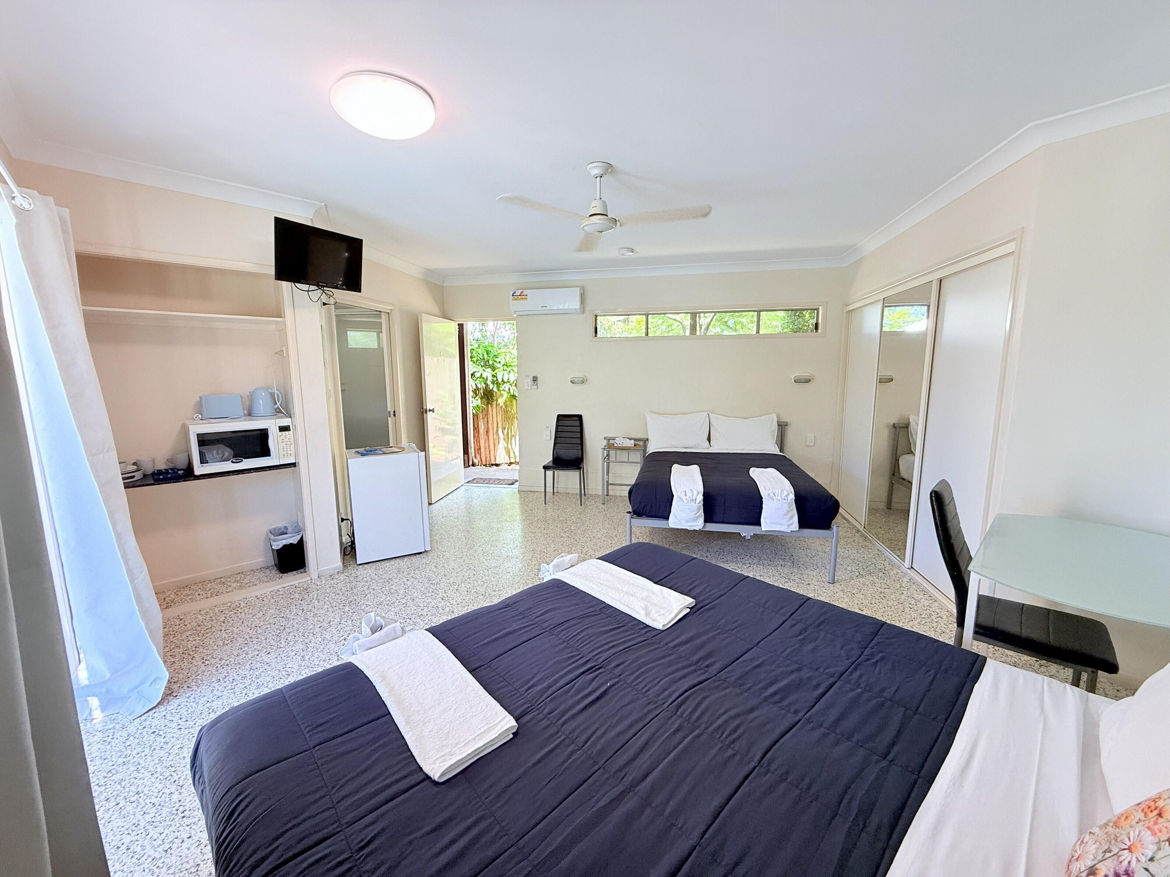 A hotel room with a bed, a desk, a chair, a TV, a ceiling fan, and a small kitchenette.