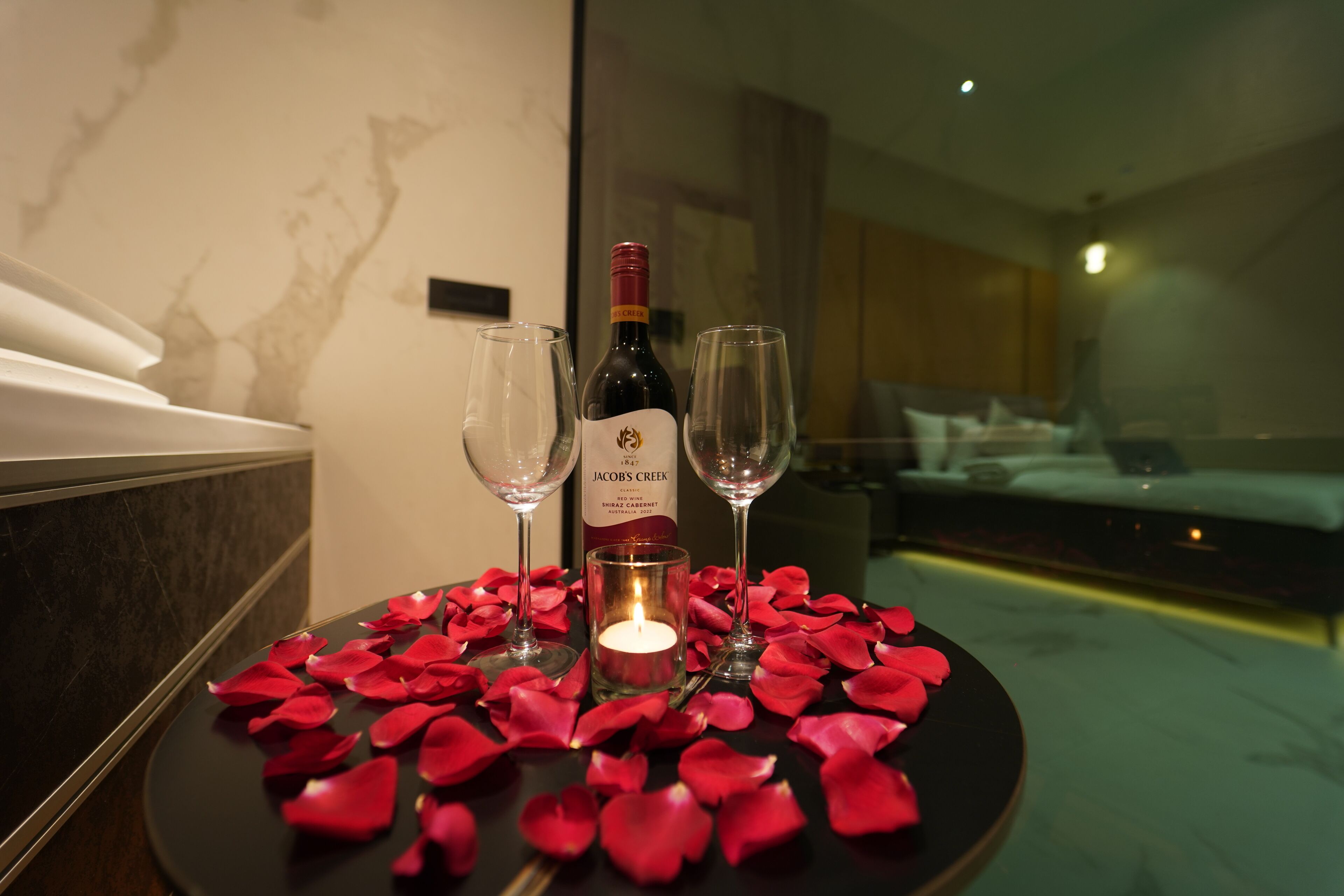 A table with rose petals, a candle, and two wine glasses with a bottle of Jacob's Creek wine.