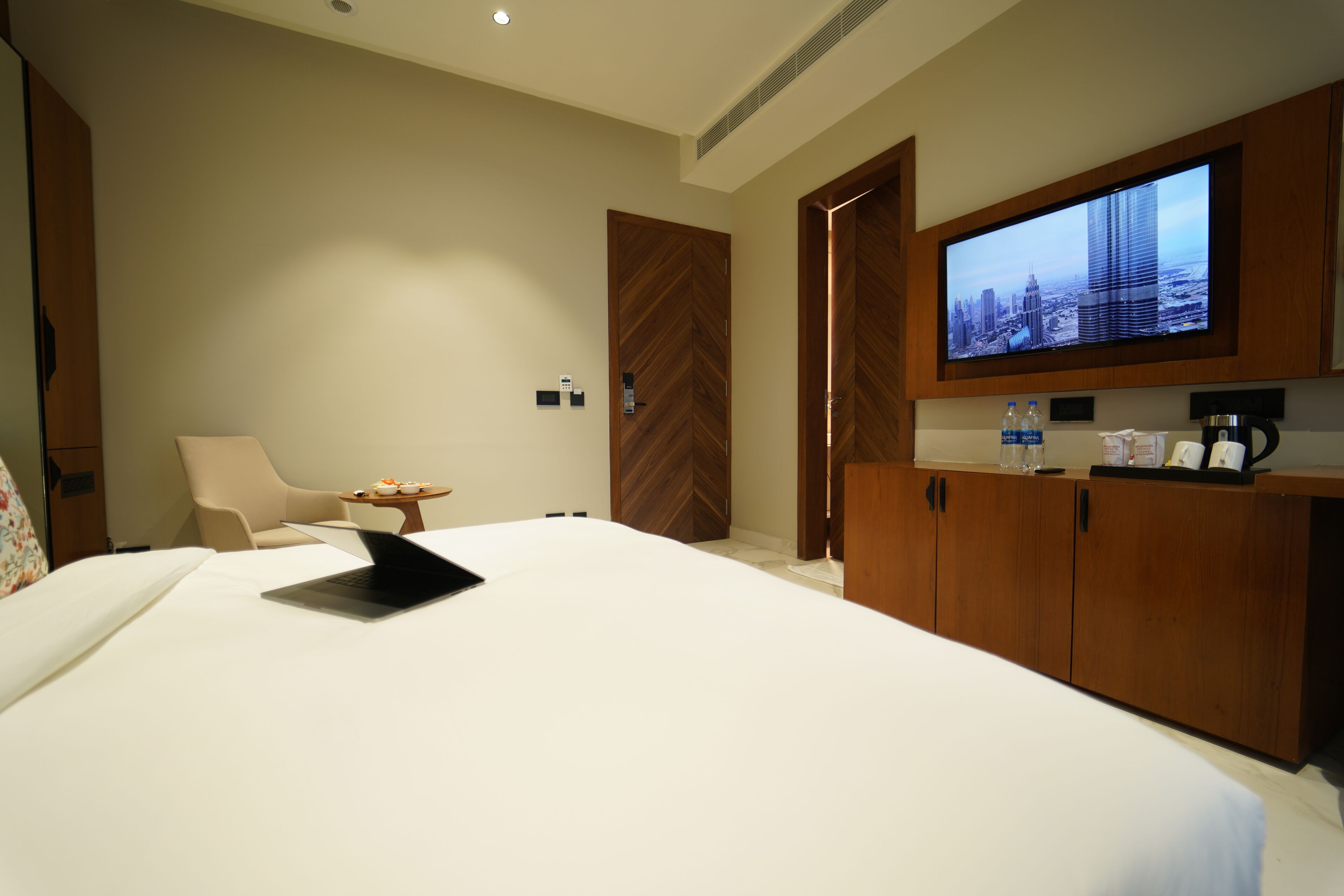 A hotel room with a bed, a laptop, a television mounted on the wall, a wooden wardrobe, and a small table with a chair.
