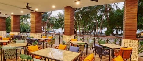 An outdoor dining area with tables and wicker chairs, surrounded by wooden pillars and palm trees.