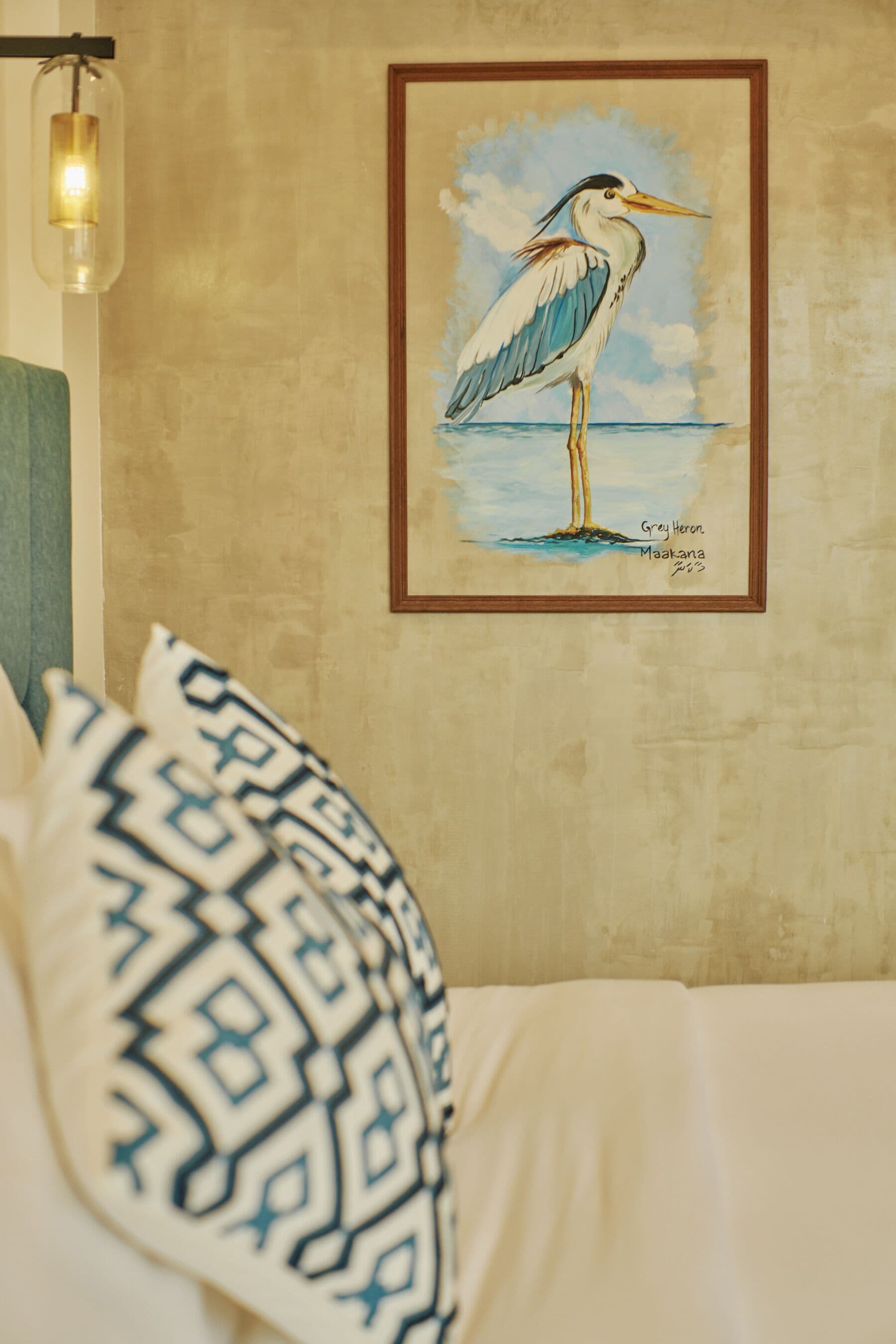A bed with patterned pillows, a framed painting of a bird, and a hanging light fixture.