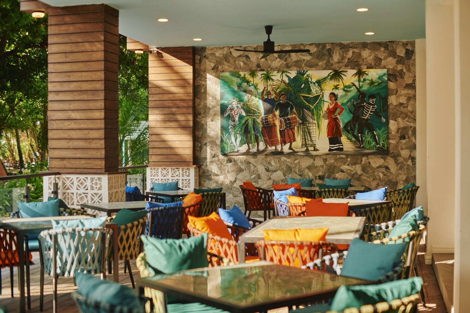 An outdoor seating area with wicker furniture and cushions, a stone wall, and a mural depicting tropical dancers.