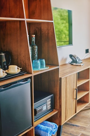 A compact kitchen with a refrigerator, a coffee maker, and a microwave. There is a wooden cabinet with a glass shelf holding a bottle and glasses. A telephone and a remote control are on the cabinet. - Beach Hotel Dhigurah (Dhigurah)