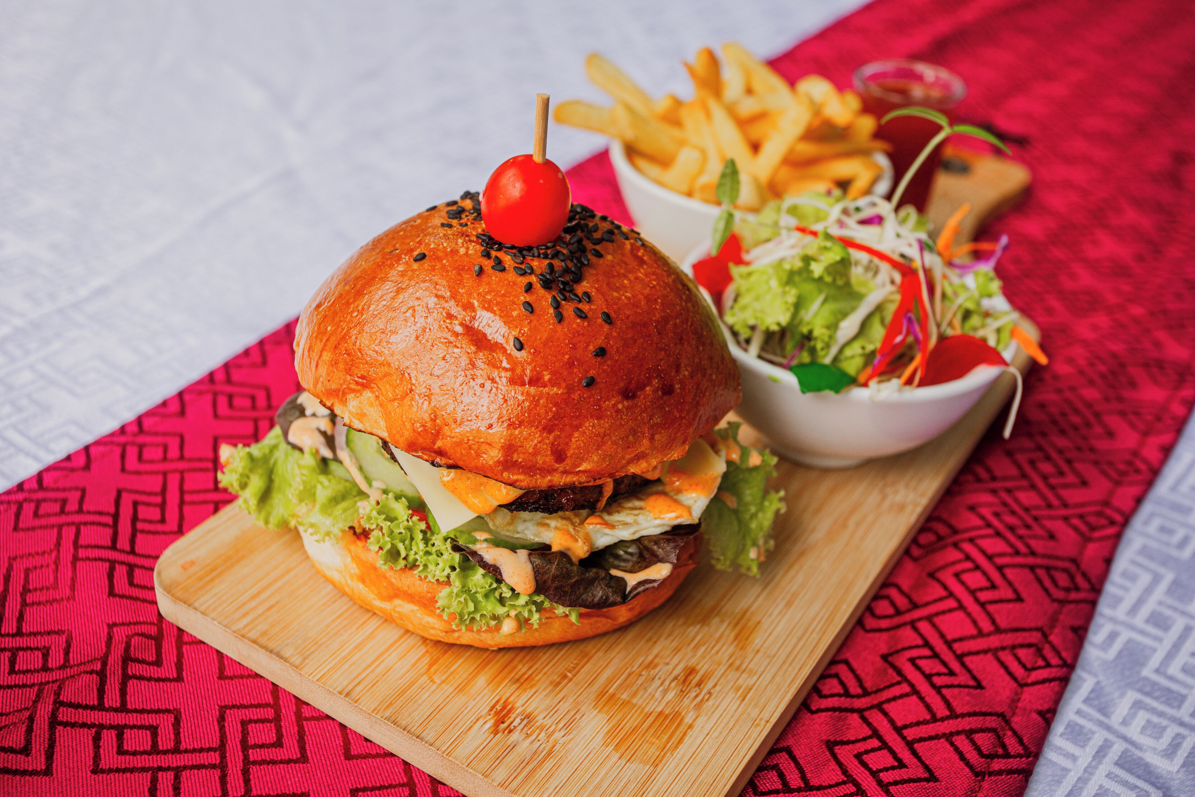 A burger with a cherry on top, a side of fries, and a salad on a wooden board.