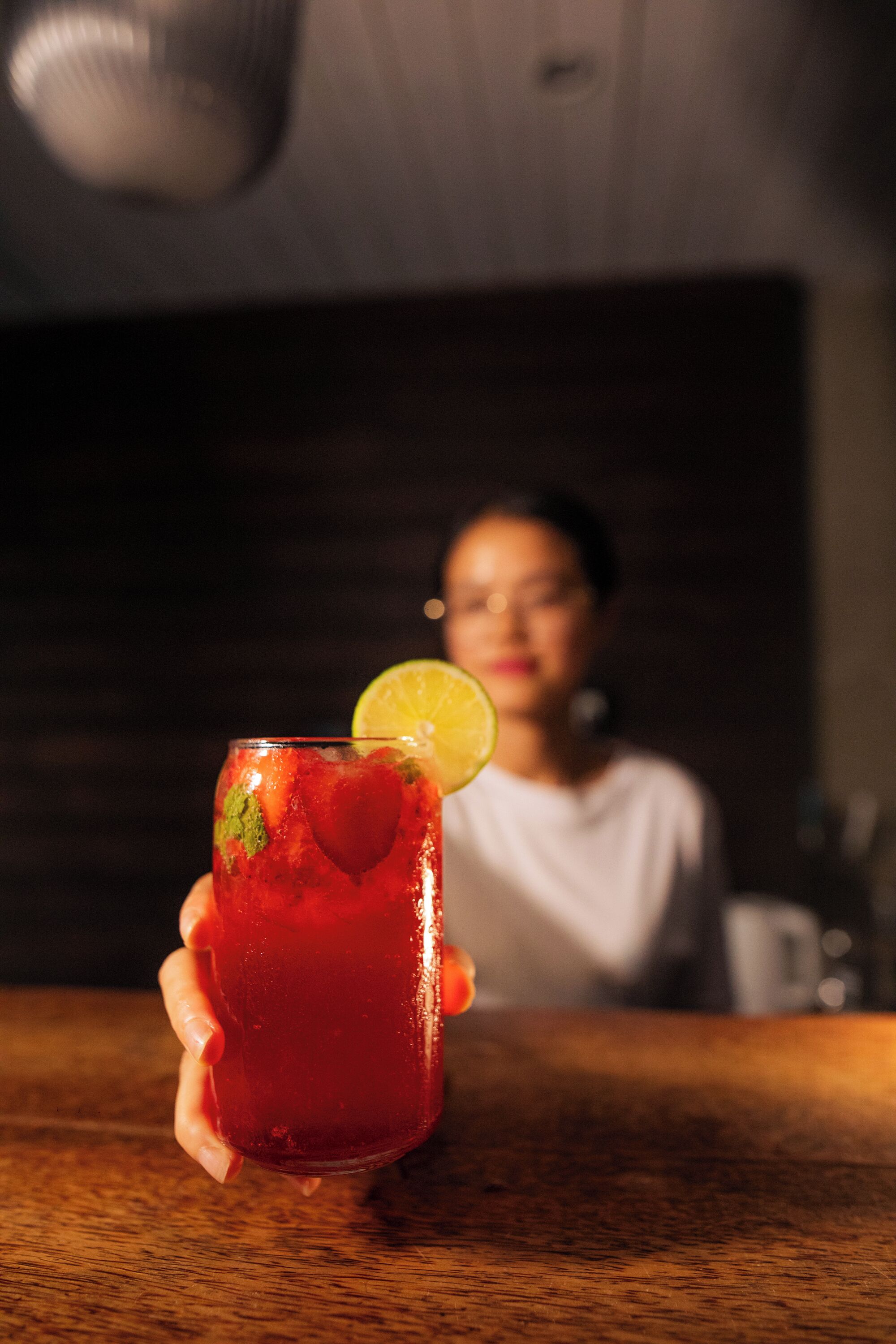 Una persona sosteniendo un vaso con una bebida roja, adornado con una rodaja de lima, en un bar con mostrador de madera.