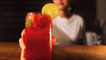 A person holding a glass with a red beverage, garnished with a slice of lime, at a bar with a wooden countertop.