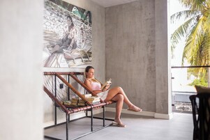 A person sitting on a hammock chair in a room with a large mural on the wall, a concrete pillar, and a view of palm trees.