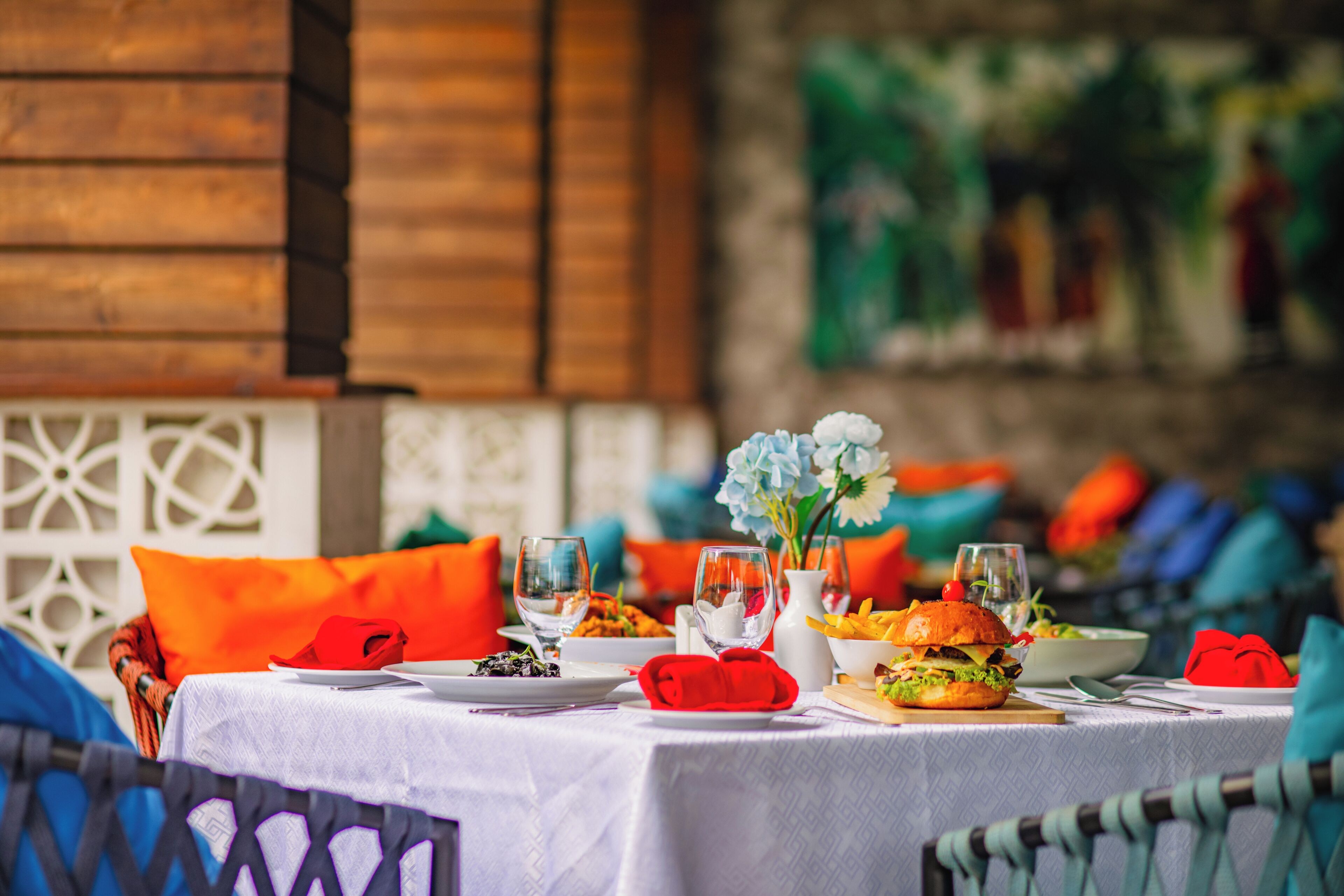 A dining table set with plates, glasses, and a burger, with a wooden wall and colorful cushions in the background.