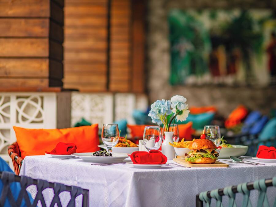 A dining table set with plates, glasses, and a burger, with a wooden wall and colorful cushions in the background.