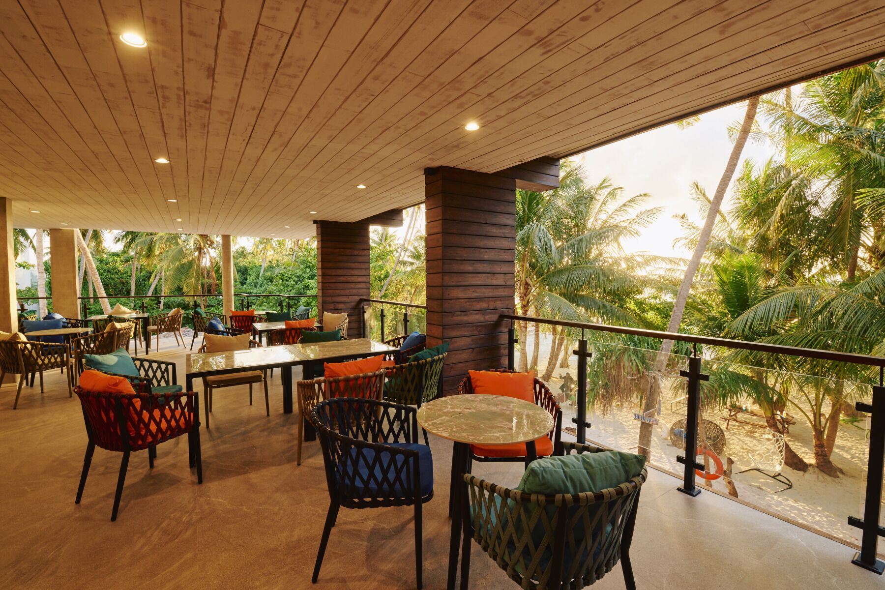 An outdoor seating area with tables and chairs, surrounded by wooden pillars and a view of palm trees.