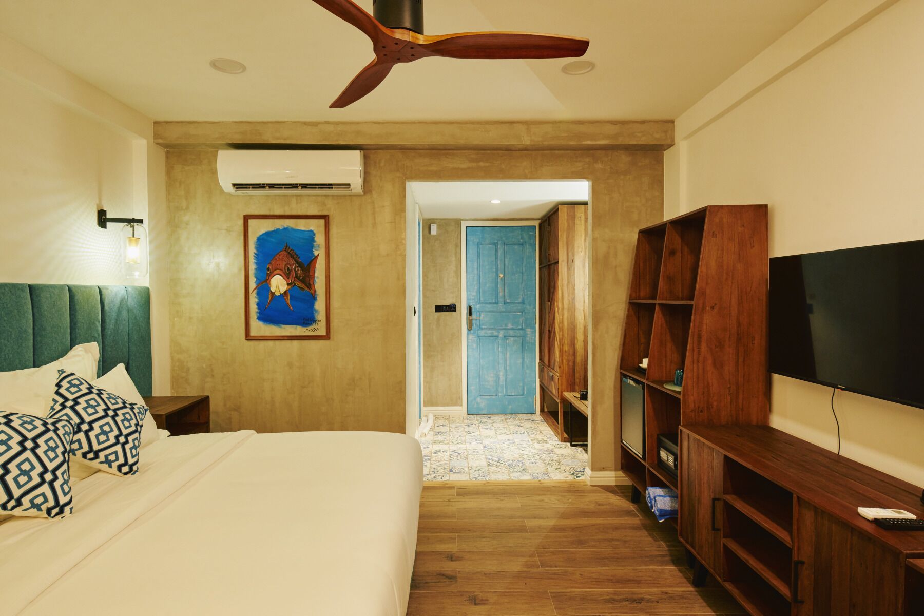 A hotel room with a bed, a ceiling fan, a TV, a wooden bookshelf, and a painting on the wall.
