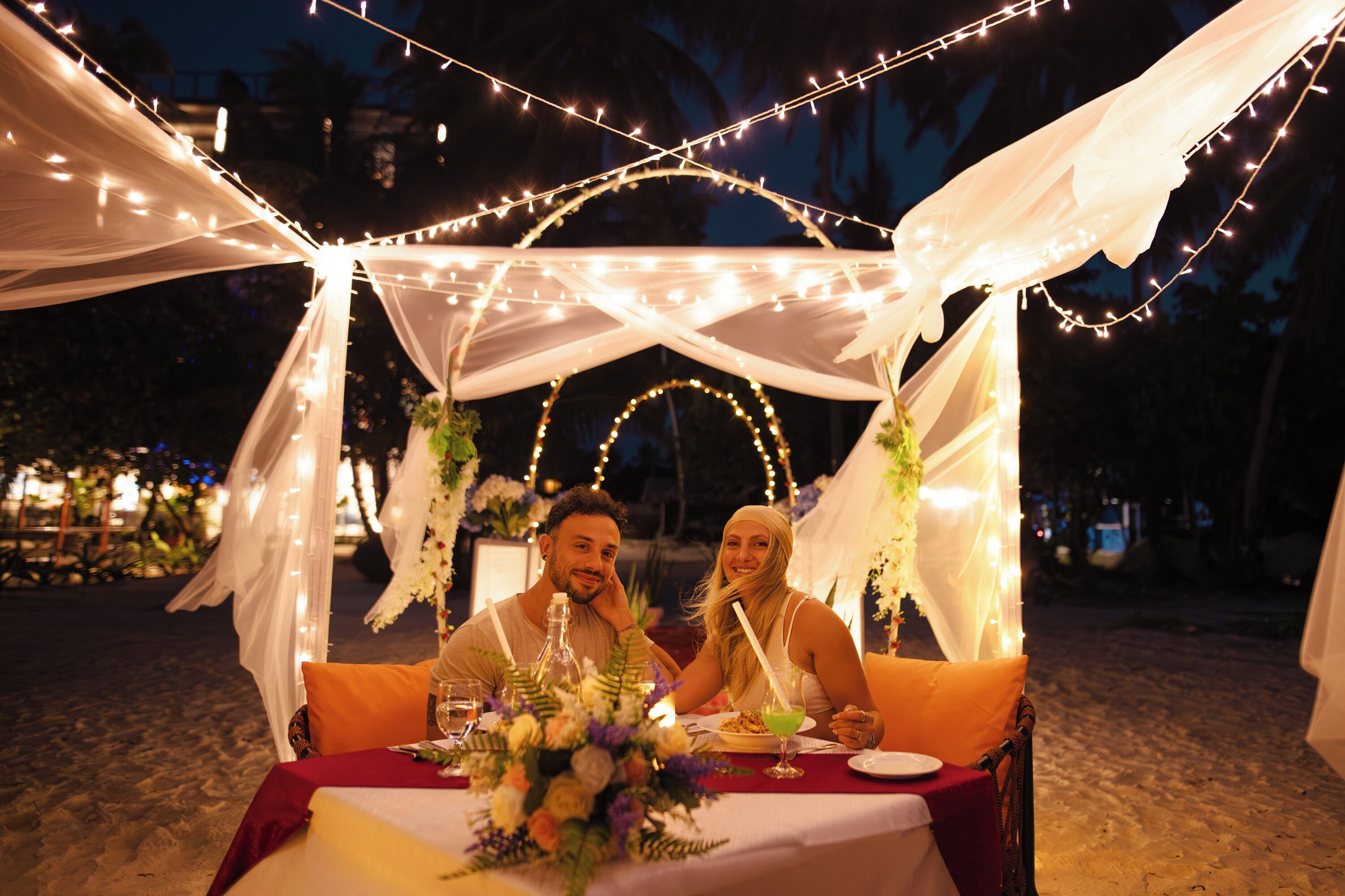A couple dining at an outdoor table with string lights and draped fabric, surrounded by greenery.