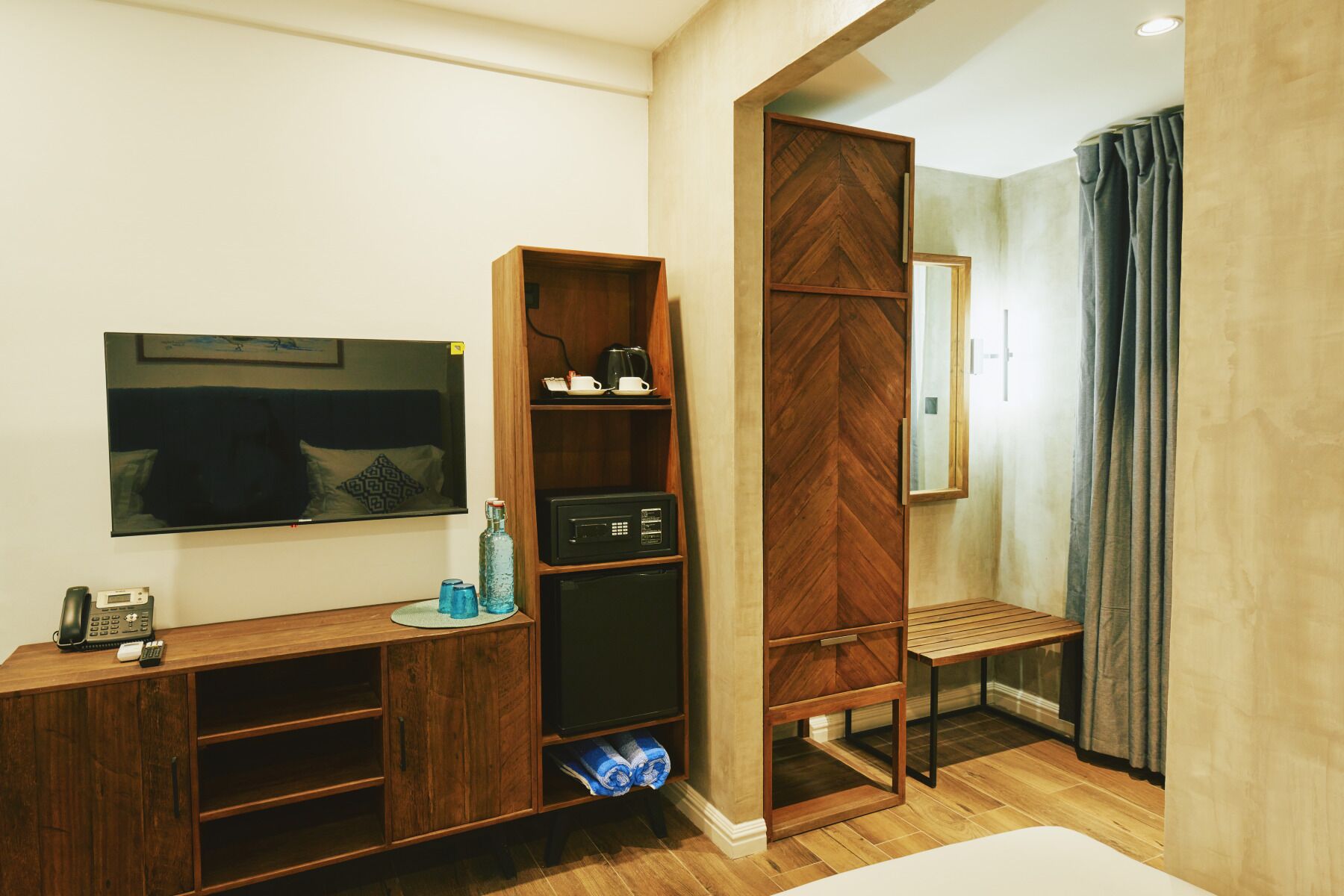 A compact hotel room with a wooden desk, a flat-screen TV, a wardrobe, and a small seating area.