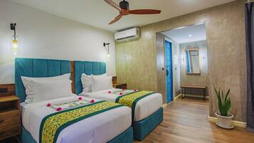 Habitación de hotel con dos camas, ventilador de techo, equipo de aire acondicionado, piso de madera, un banco y una planta en maceta.