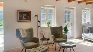 A living room with two armchairs, a small ottoman, a floor lamp, and a painting on the wall. There are three windows providing natural light.