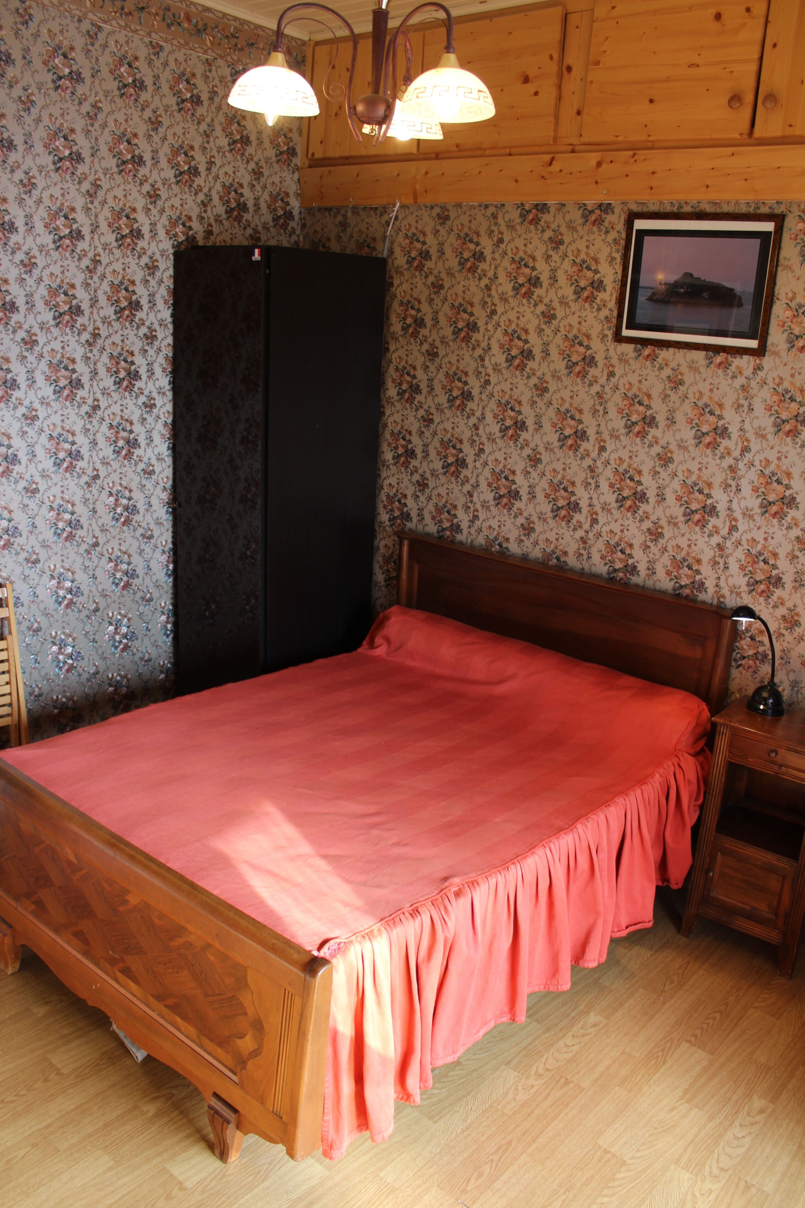 A bedroom with a wooden bed, floral wallpaper, a black wardrobe, and a framed picture on the wall.