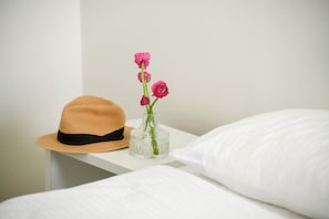 A bed with white bedding, a white nightstand with a straw hat and a clear vase with pink flowers.