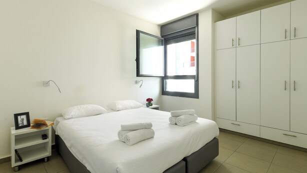 A neatly made bed with white linens and towels, a bedside table with a picture frame and books, a nightstand with a lamp, and a wardrobe with multiple doors. - 1 BR Apt w living room & learning area (Tel Aviv)