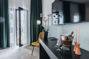 A modern hotel room with a large window, a yellow chair, a black desk, a potted orchid, a champagne bucket, and pink candle holders.