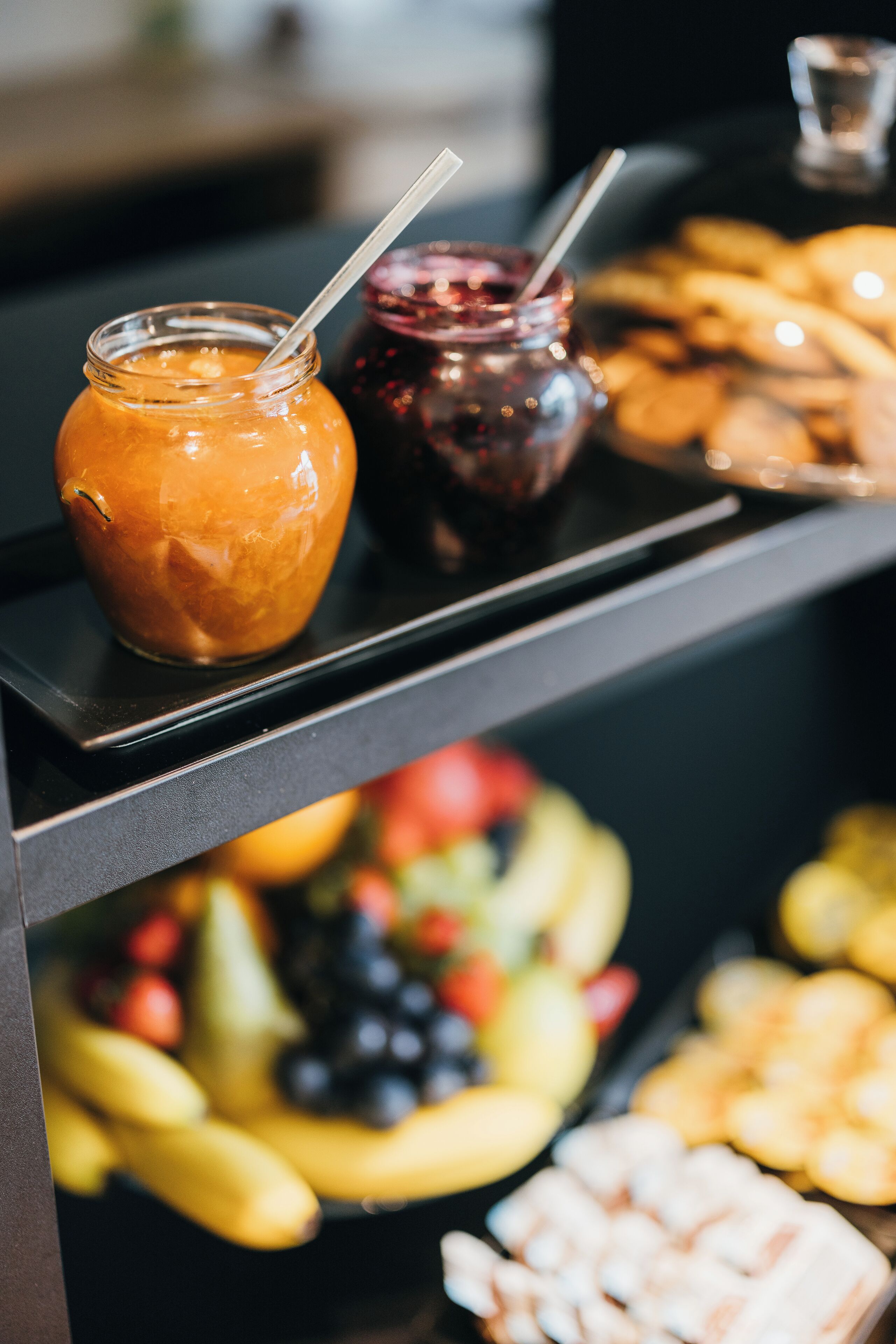 A buffet table with jars of orange and red sauces, a tray of cookies, and a variety of fruits including bananas, apples, and grapes.