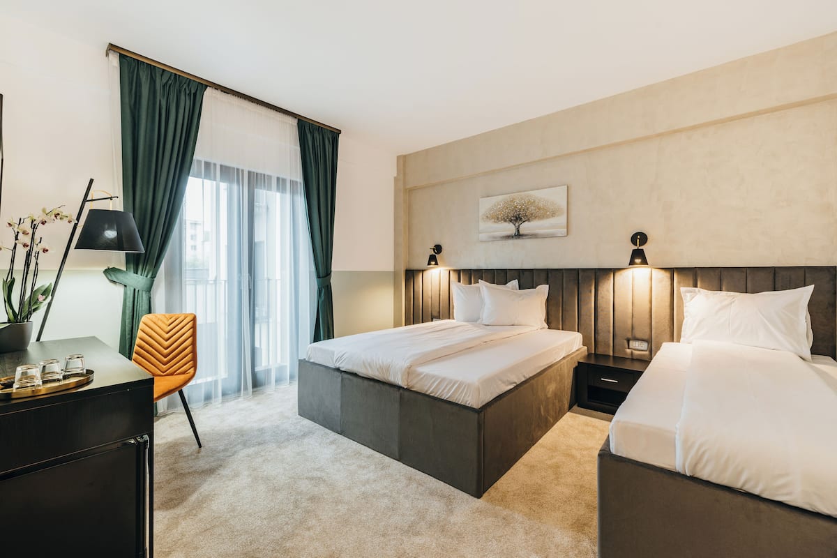 A modern hotel room with a large bed, a desk with a chair, a nightstand, and a wall-mounted lamp.