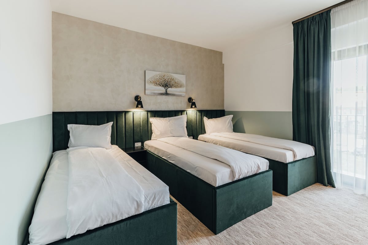 A hotel room with two single beds, a large window with curtains, a wall-mounted painting, and two wall sconces.