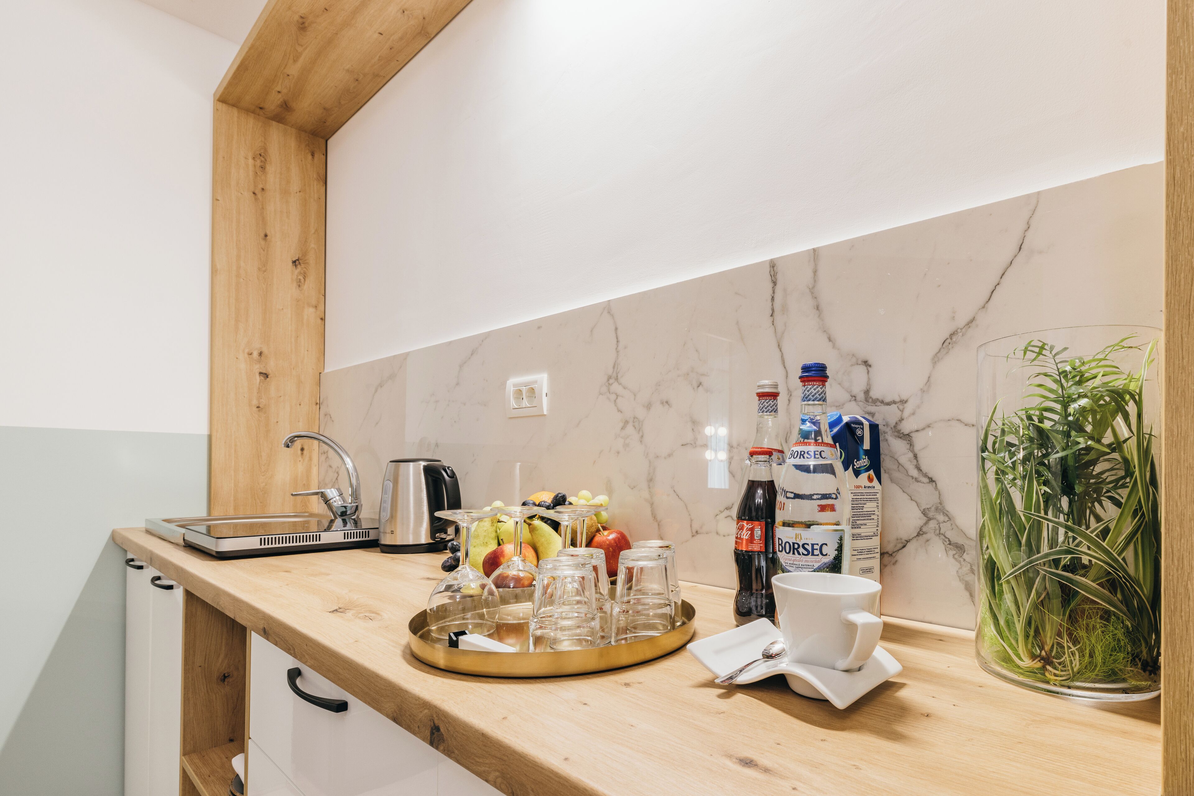 Une cuisine avec un plan de travail en bois, un pare-feu en marbre et une machine à café. On y trouve des fruits, des verres et une bouteille de Borsec sur le plan de travail.