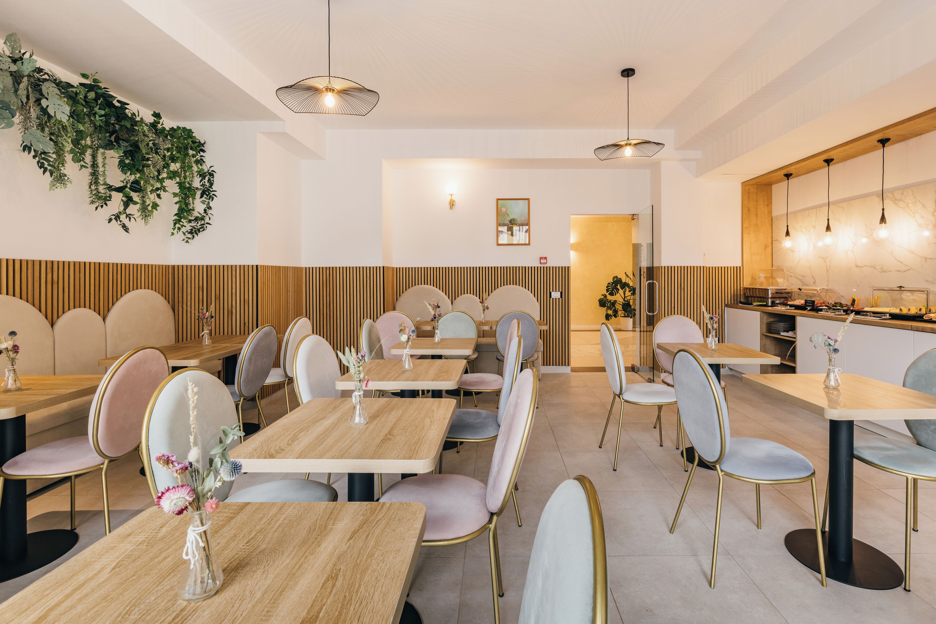 Interior moderno de restaurante con mesas y sillas de madera, iluminación colgante y una zona de cocina visible al fondo.