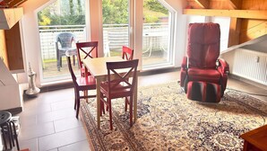 A cozy attic room with a wooden ceiling, a red massage chair, a dining table with chairs, and large windows offering a view of the backyard. - Apartment Kelkheim (Kelkheim (Taunus))
