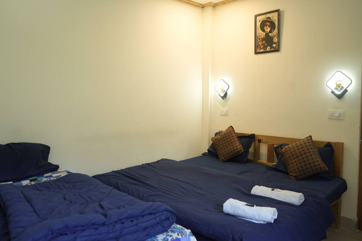 A hotel room with two beds, each with dark blue bedding and multiple pillows. There are wall-mounted light fixtures on either side of the beds.