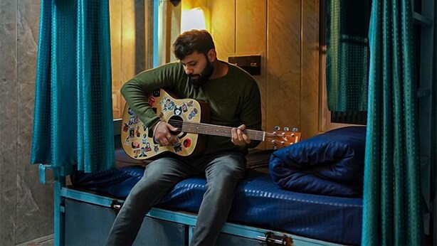 Une personne joue de la guitare dans une chambre avec des lits superposés, dont l’intérieur est en bois et le matelas est bleu.