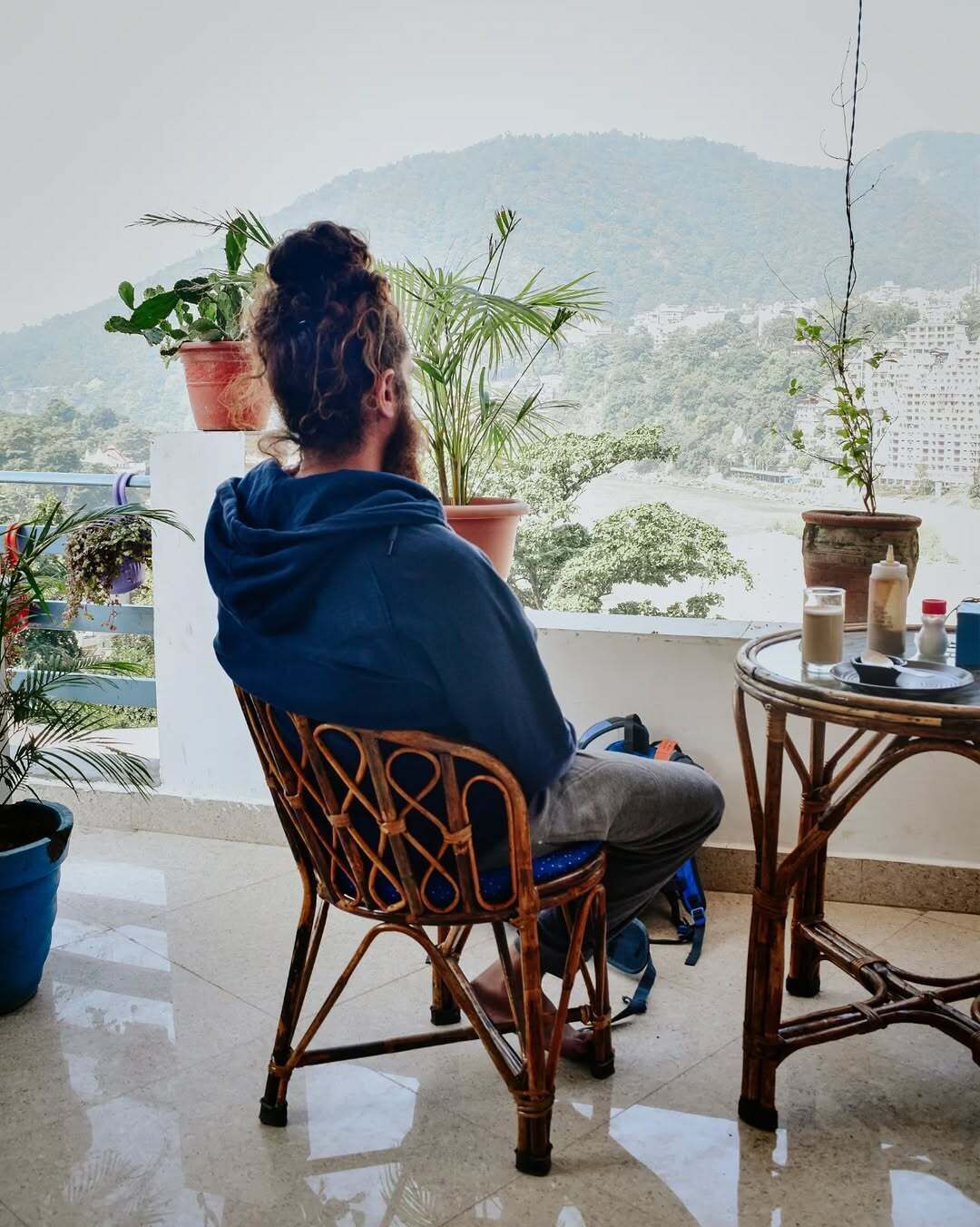 Une personne assise sur une chaise en osier, sur un balcon agrémenté de plantes en pot, avec vue sur le paysage urbain.