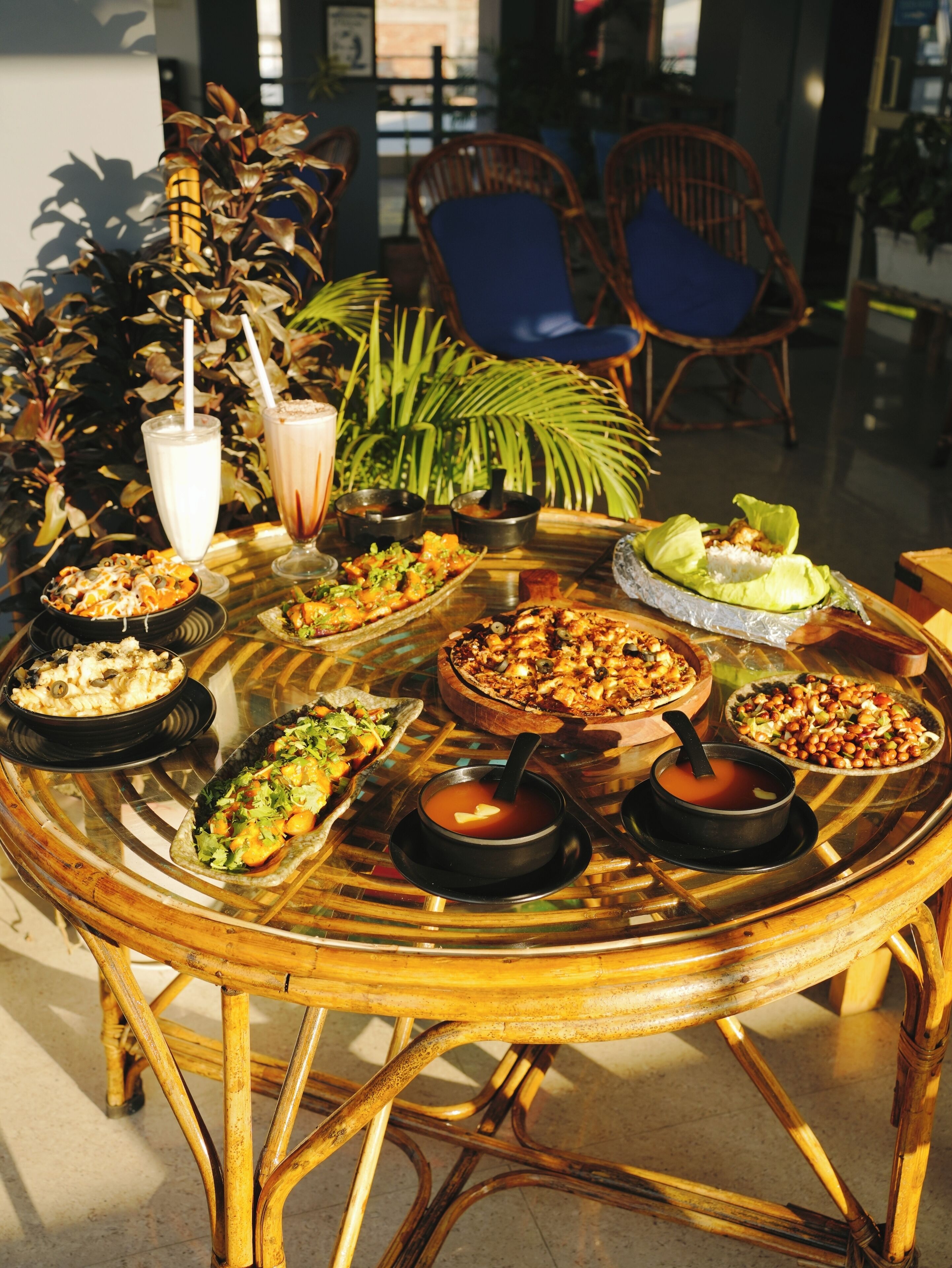 A round glass table with various dishes and drinks, surrounded by wicker chairs and potted plants.